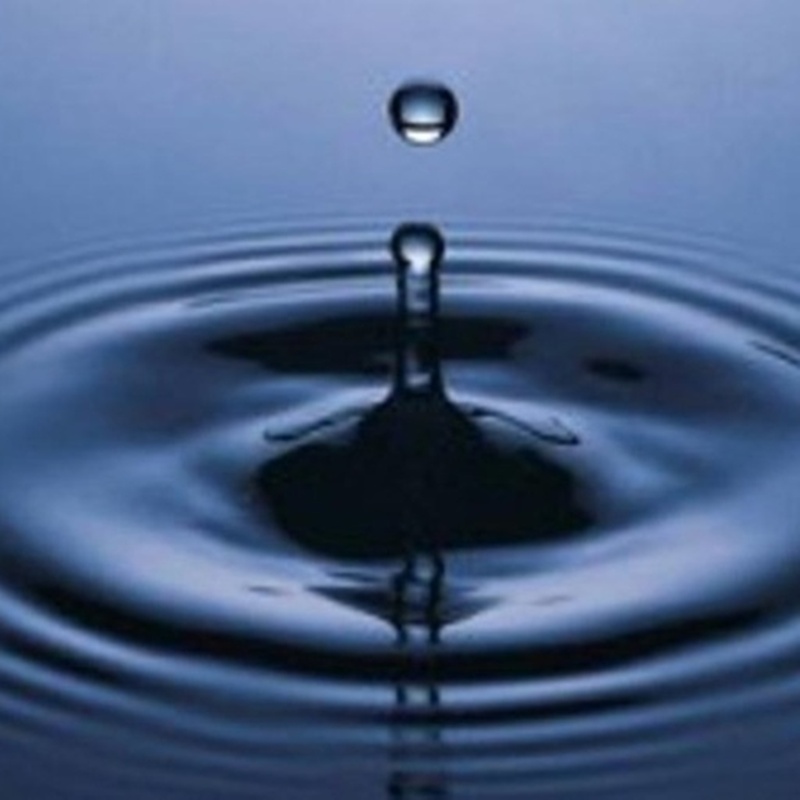 Close-up view of a clean water droplet striking a calm blue surface, creating perfect concentric rippling waves.
